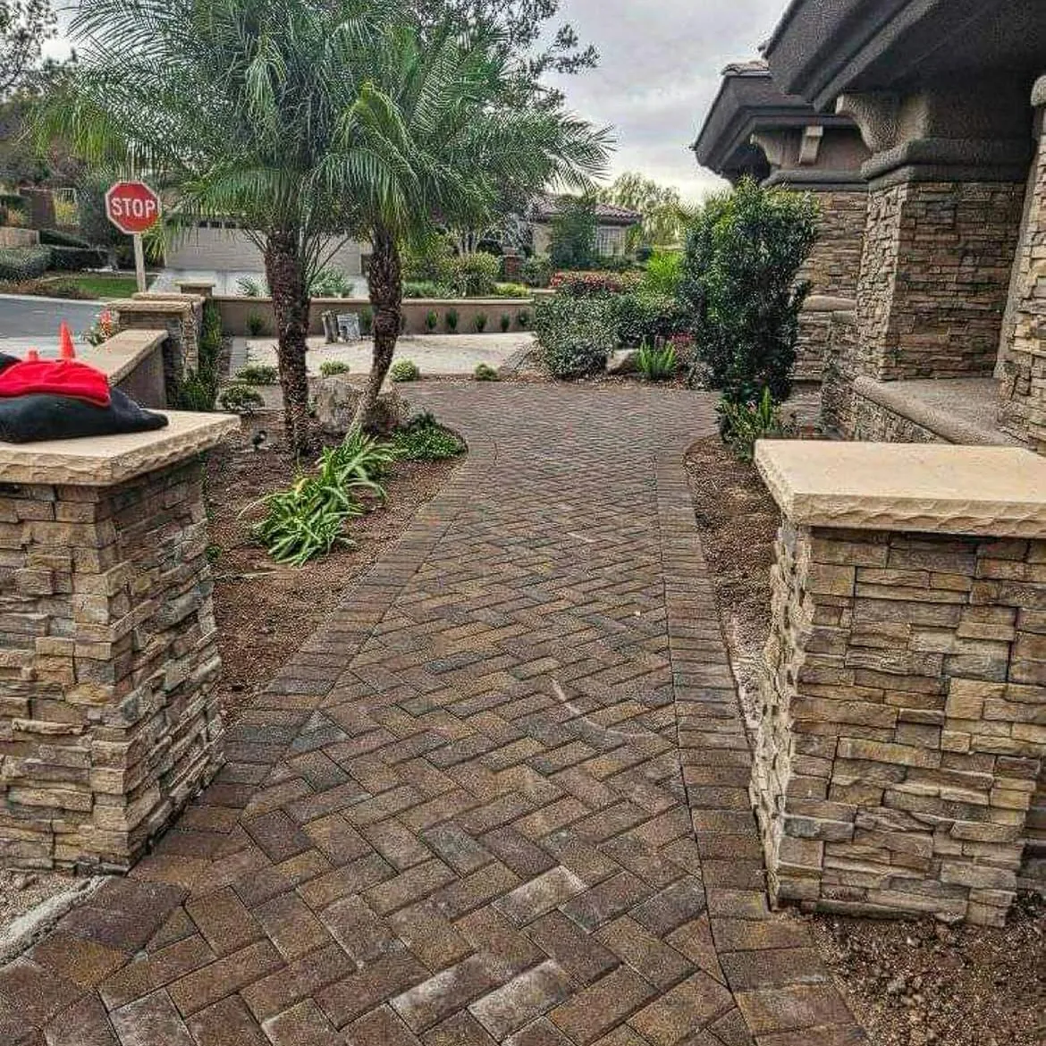 Herringbone paver drive between stacked-stone pillars, with palms and shrubs.