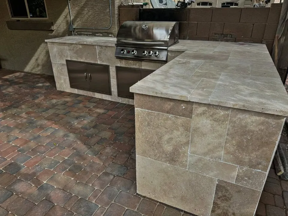 L-shaped travertine outdoor kitchen with a built-in grill on a brick paver patio.